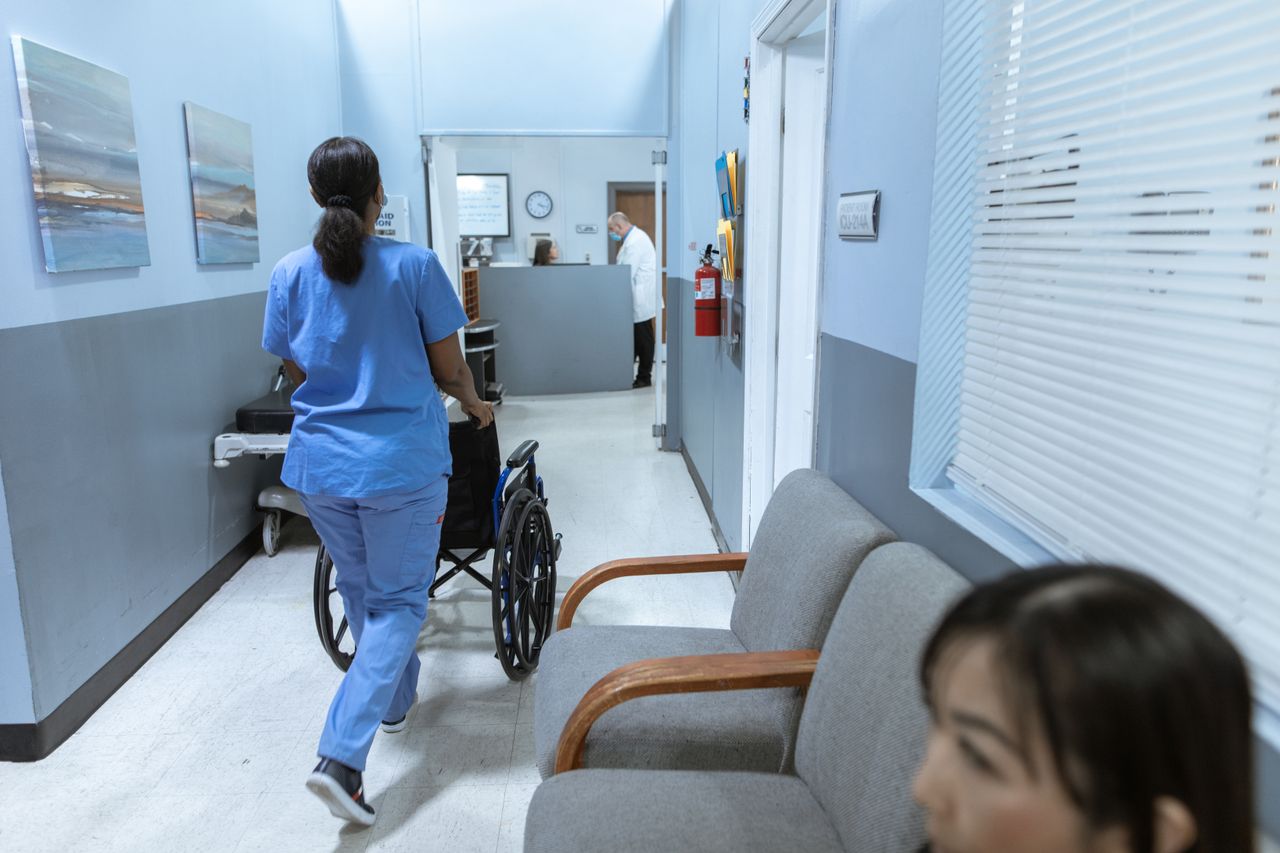 Home A nurse in scrubs pushes a wheelchair through a well-lit hospital corridor, attending to patient care.