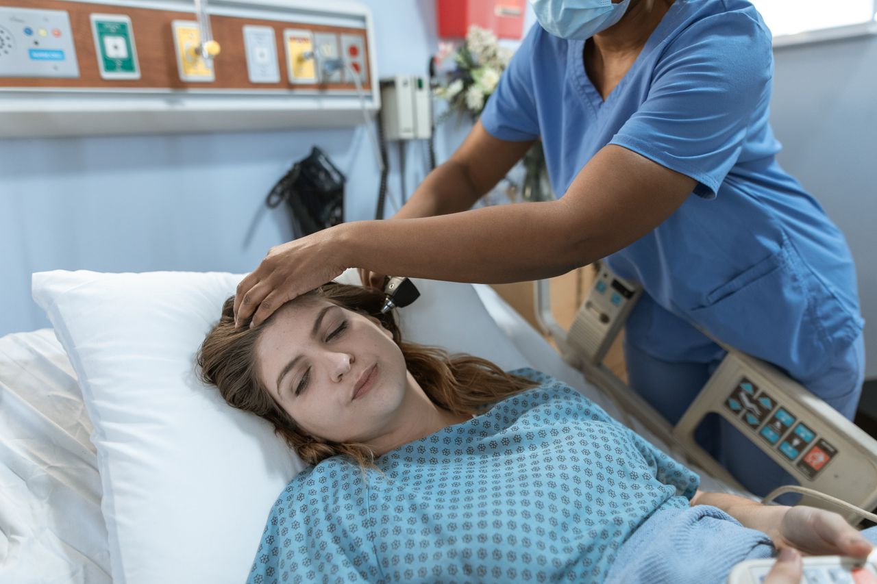 Home Crop African American female doctor with professional equipment doing examination of ear of woman lying on bed in hospital ward